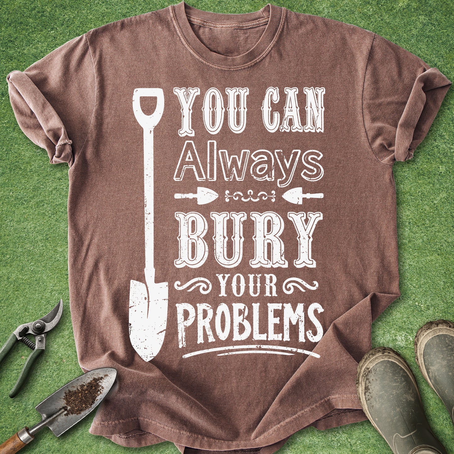 A brown short-sleeve graphic T-shirt laid flat on green grass-like ground, with gardening tools and boots around it. The shirt has a large white distressed text print reading “YOU CAN Always BURY YOUR PROBLEMS” with a shovel illustration.