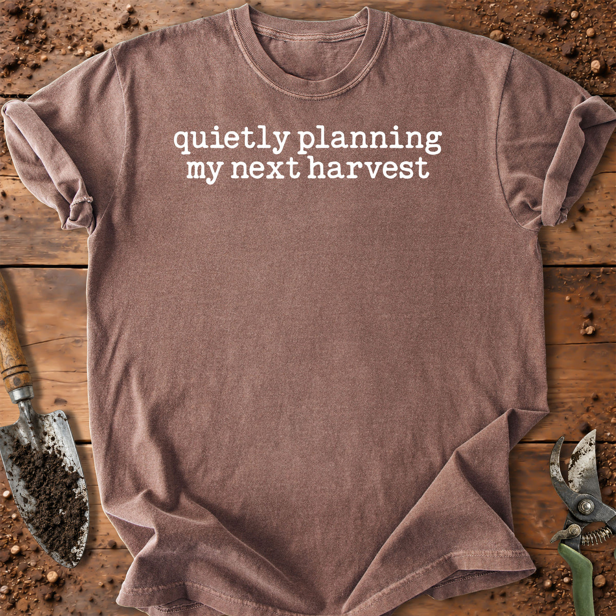 A brown short-sleeve t-shirt with the white printed text 'quietly planning my next harvest' laid flat on a rustic wooden surface with gardening tools and soil.