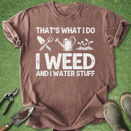A brown short-sleeve graphic T-shirt laid flat on green artificial grass, with gardening tools and boots around it. The shirt has a white distressed slogan reading “THAT’S WHAT I DO I WEED AND I WATER STUFF” with small gardening icons above the text.
