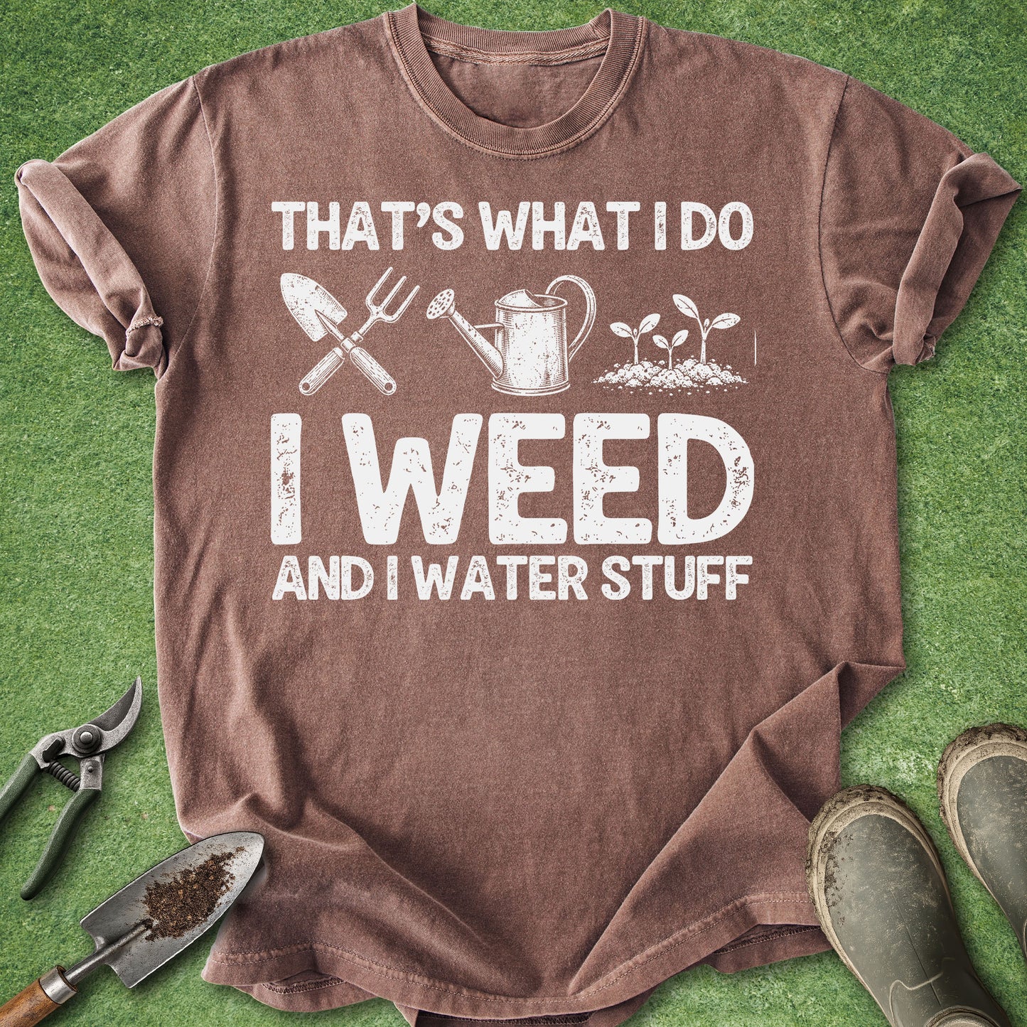 A brown short-sleeve graphic T-shirt laid flat on green grass-like background, with gardening tools and boots around it. The shirt has large white text reading 'THAT'S WHAT I DO I WEED AND I WATER STUFF' with simple white illustrations of a trowel, fork, watering can, and seedlings.