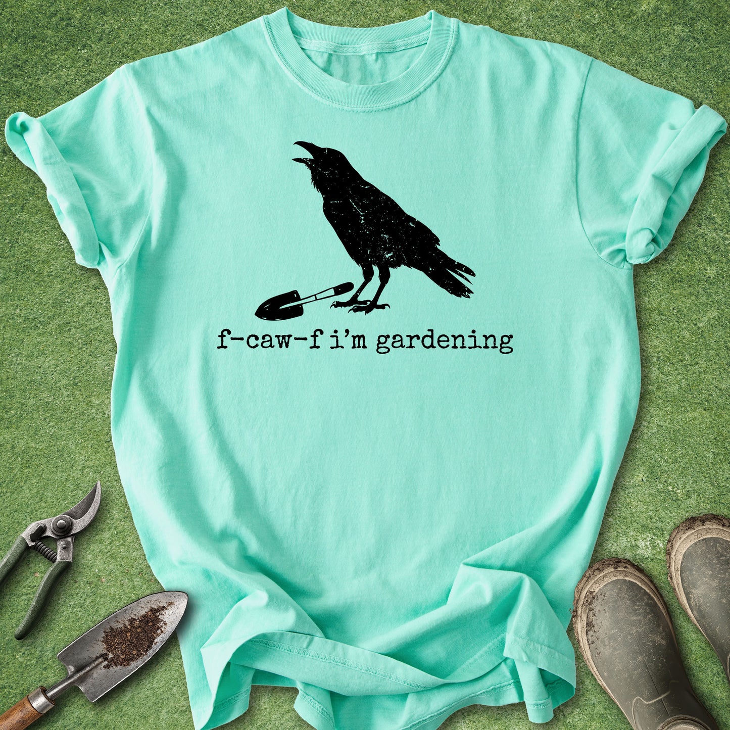 A mint green graphic t-shirt laid flat on a green grass-like surface, surrounded by gardening props including pruning shears, a trowel, and muddy boots. The shirt features a black graphic of a crow holding a small shovel, with the text 'f-caw-f i'm gardening' printed below.