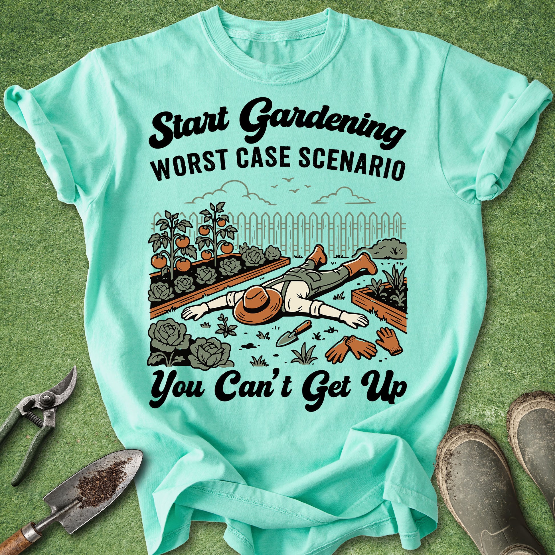 A mint green graphic t-shirt laid flat on a green grass-like surface, surrounded by gardening props including pruning shears, a trowel, gloves, and boots. The shirt features a humorous illustration of a gardener lying on the ground with the text 'Start Gardening WORST CASE SCENARIO You Can't Get Up'.