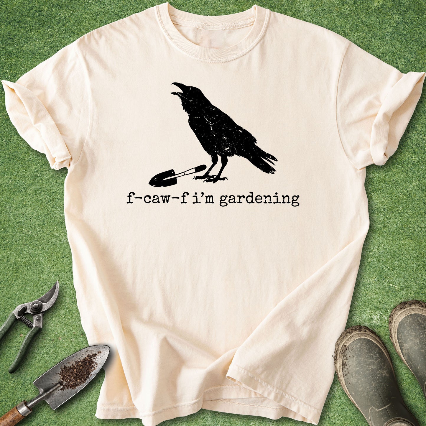 A cream-colored short-sleeve t-shirt with a black graphic of a crow standing on a garden fork and the text 'f-caw-f i'm gardening'. The shirt is laid flat on a green grass-like surface, surrounded by gardening props including pruning shears, a small hand trowel, and muddy boots.
