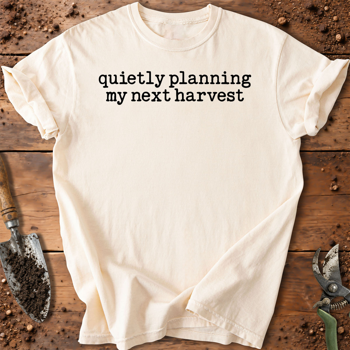 An off-white short-sleeve t-shirt with the black printed text 'quietly planning my next harvest' laid flat on a rustic wooden surface with gardening tools.