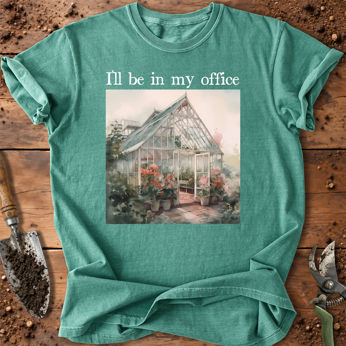 A teal green graphic t-shirt laid flat on a rustic wooden surface with gardening tools. The shirt features a printed illustration of a greenhouse surrounded by flowers and the text 'I'll be in my office' in white above the graphic.