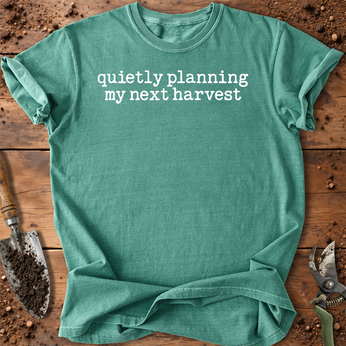 A teal green short-sleeve t-shirt with the white printed text 'quietly planning my next harvest' laid flat on a rustic wooden surface with gardening tools and soil.