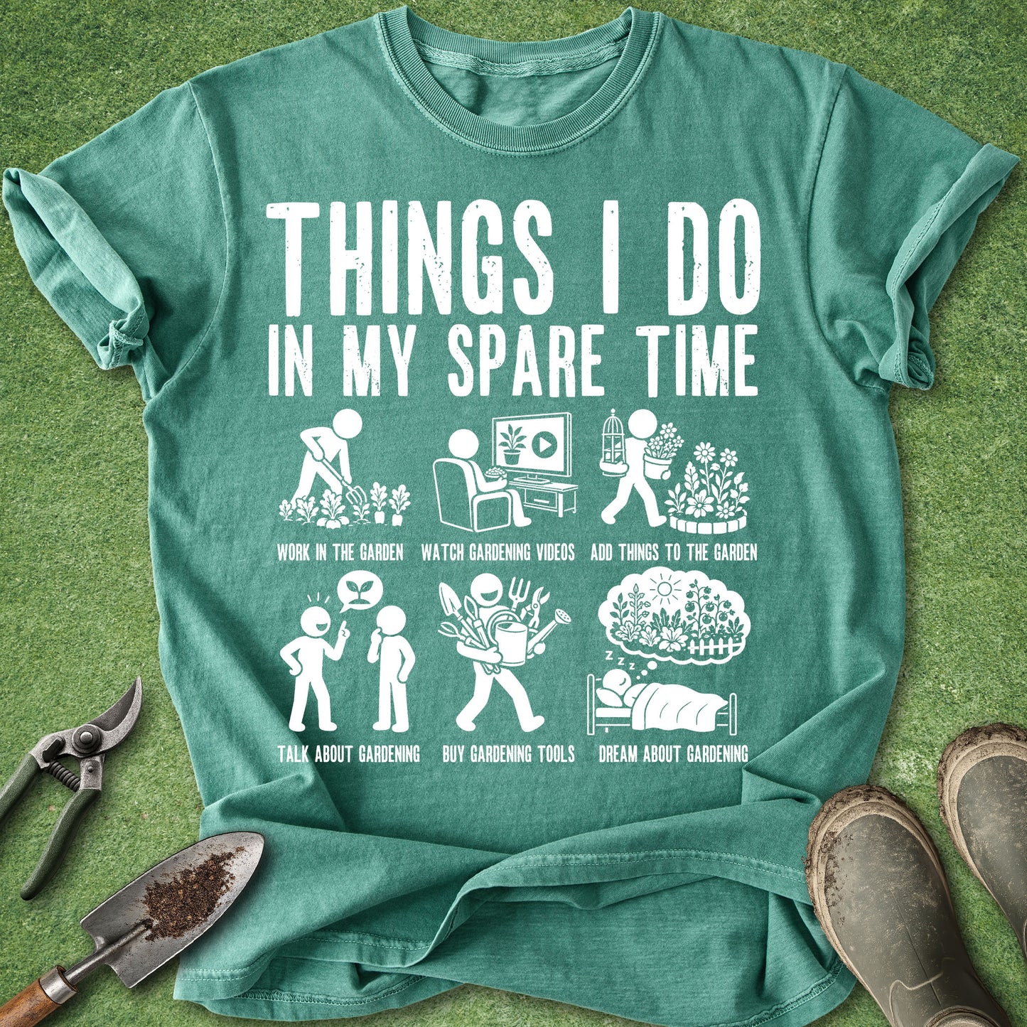 A teal green graphic t-shirt laid flat on a green grass-like surface, surrounded by gardening props including pruning shears, a trowel, and boots. The shirt features a white printed graphic titled 'THINGS I DO IN MY SPARE TIME' with six humorous gardening-themed illustrations and captions.