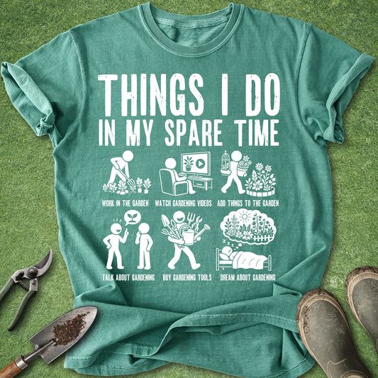 A teal green graphic t-shirt laid flat on a green grass-like surface, surrounded by gardening props including pruning shears, a trowel, and boots. The shirt features a white printed graphic titled 'THINGS I DO IN MY SPARE TIME' with six humorous gardening-themed illustrations and captions.