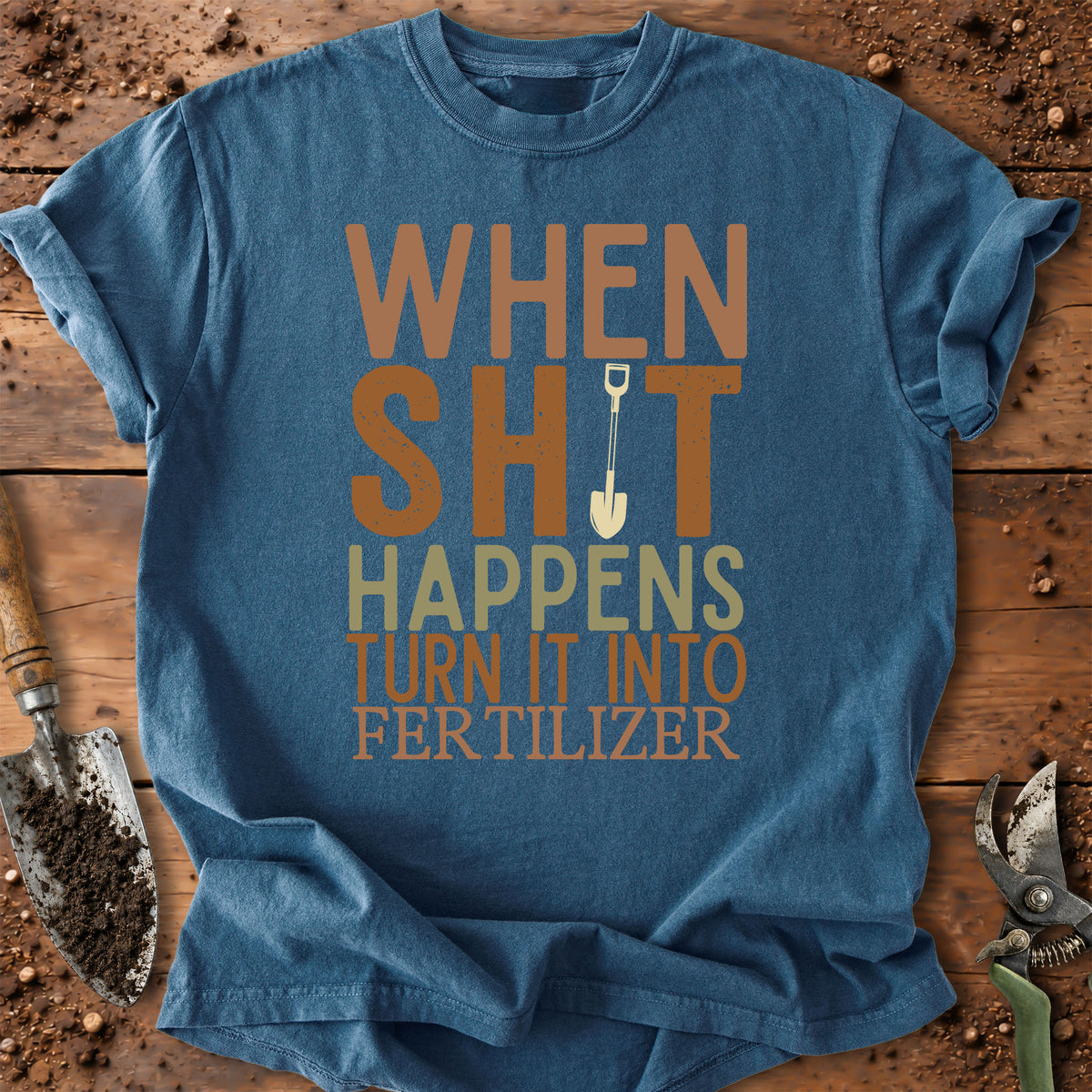 A blue graphic t-shirt laid flat on a rustic wooden surface with gardening props like a trowel, soil, and pruning shears. The shirt features a humorous graphic that reads 'WHEN SHIT HAPPENS TURN IT INTO FERTILIZER' in orange, beige, and light green lettering, with a small shovel illustration.