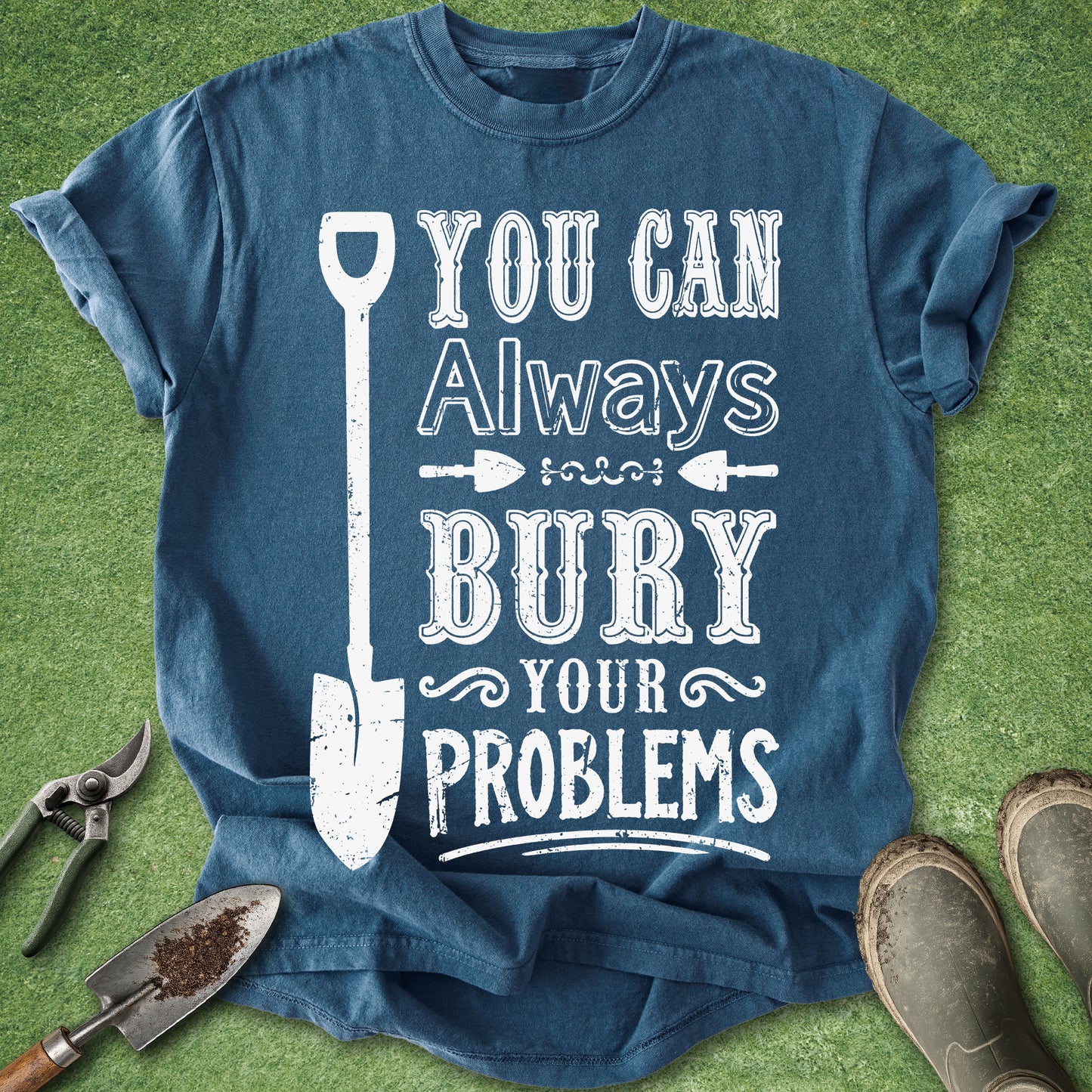 A blue graphic t-shirt laid flat on green grass with gardening tools and boots around it. The shirt has a distressed white graphic that reads 'YOU CAN Always BURY YOUR PROBLEMS' with a shovel illustration.