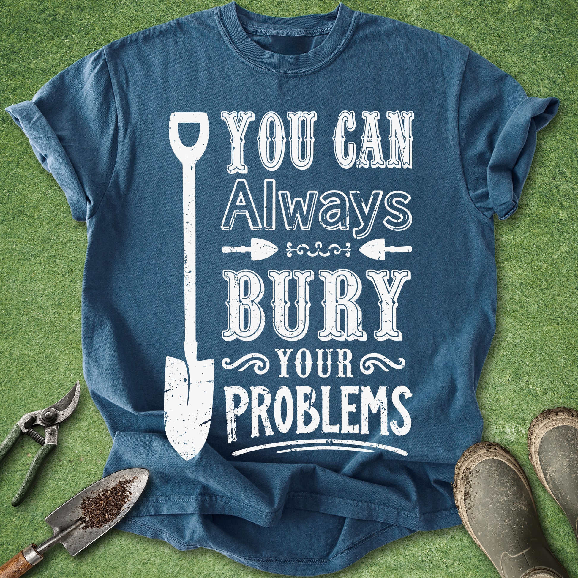 A blue graphic t-shirt laid flat on green grass with gardening tools and boots around it. The shirt has a distressed white graphic that reads 'YOU CAN Always BURY YOUR PROBLEMS' with a shovel illustration.