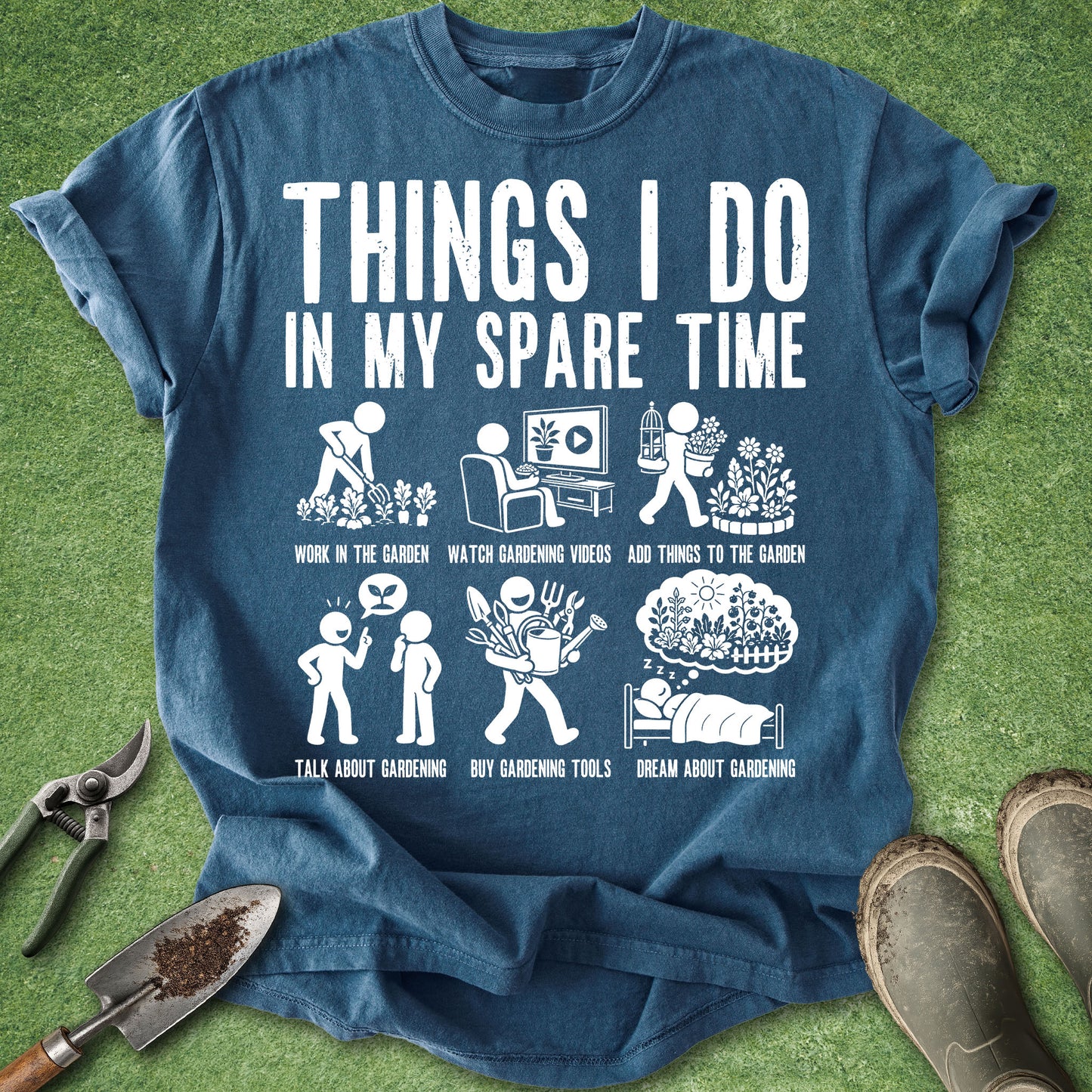 A blue graphic t-shirt laid flat on green grass with gardening props around it. The shirt has a large white printed slogan reading 'THINGS I DO IN MY SPARE TIME' above six small gardening-themed illustrations and text.