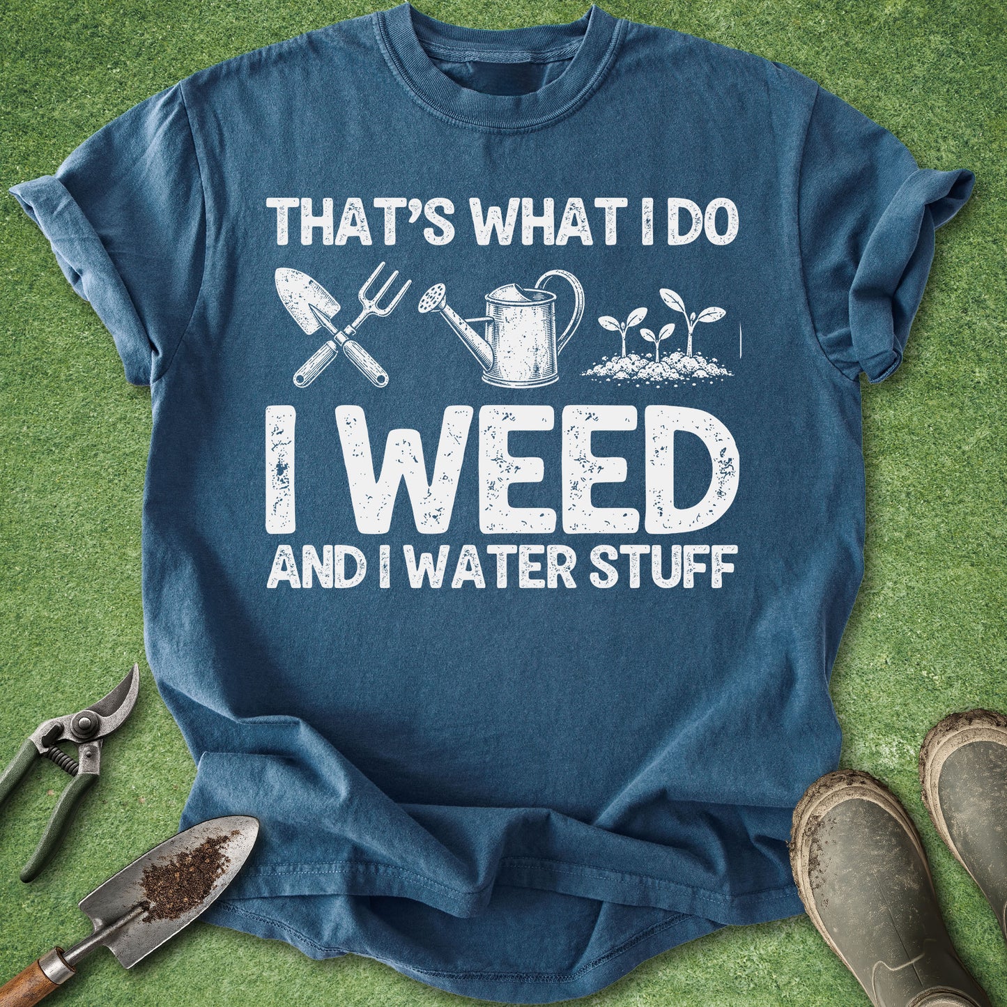 A blue graphic t-shirt laid flat on green grass with gardening props, including pruning shears, a trowel, a watering can, and boots. The shirt features white distressed text that reads 'THAT'S WHAT I DO I WEED AND I WATER STUFF' with simple gardening tool and plant illustrations.