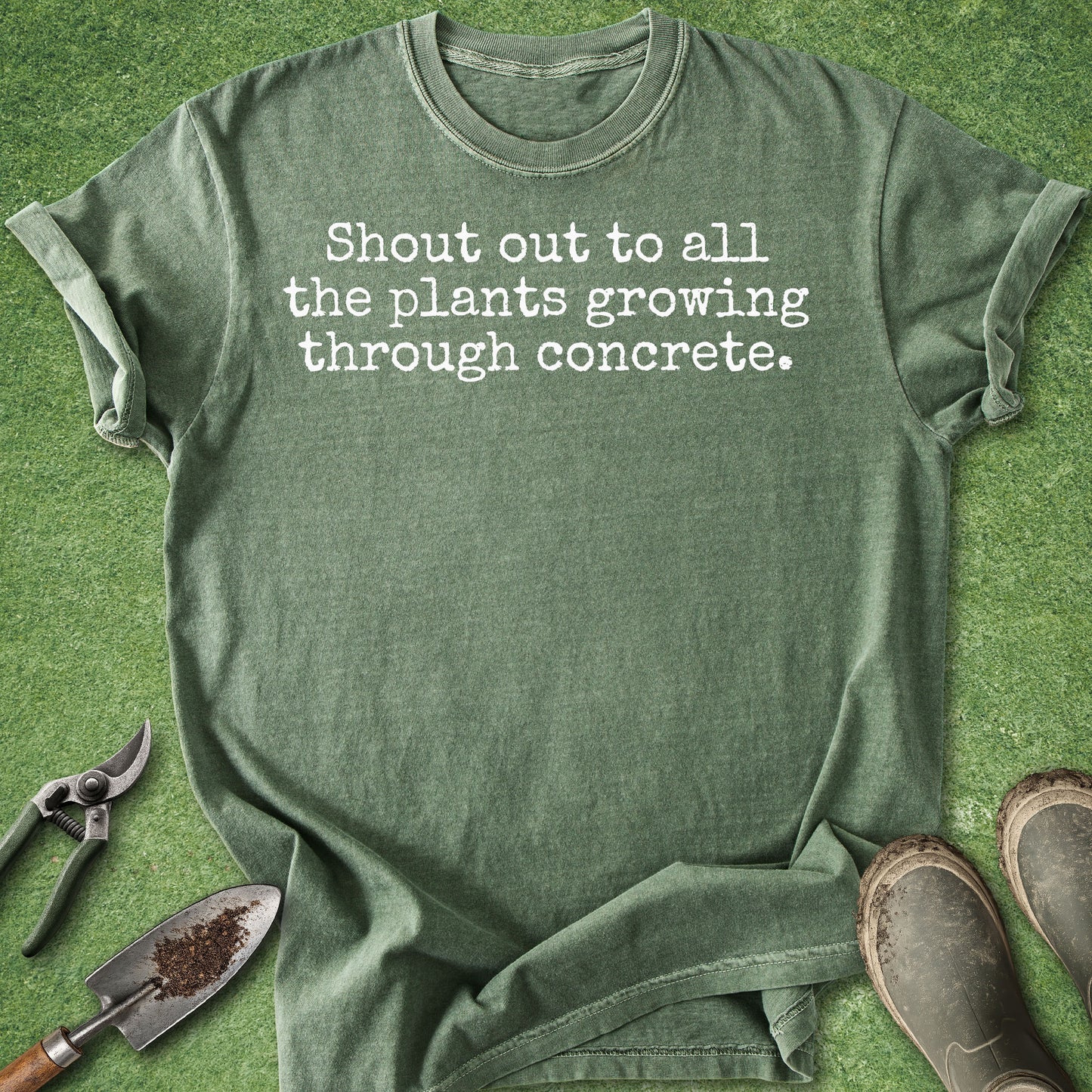 An olive green graphic t-shirt with the text 'Shout out to all the plants growing through concrete.' printed in white, styled in a flat-lay on green grass with gardening tools (trowel, pruning shears) and boots.