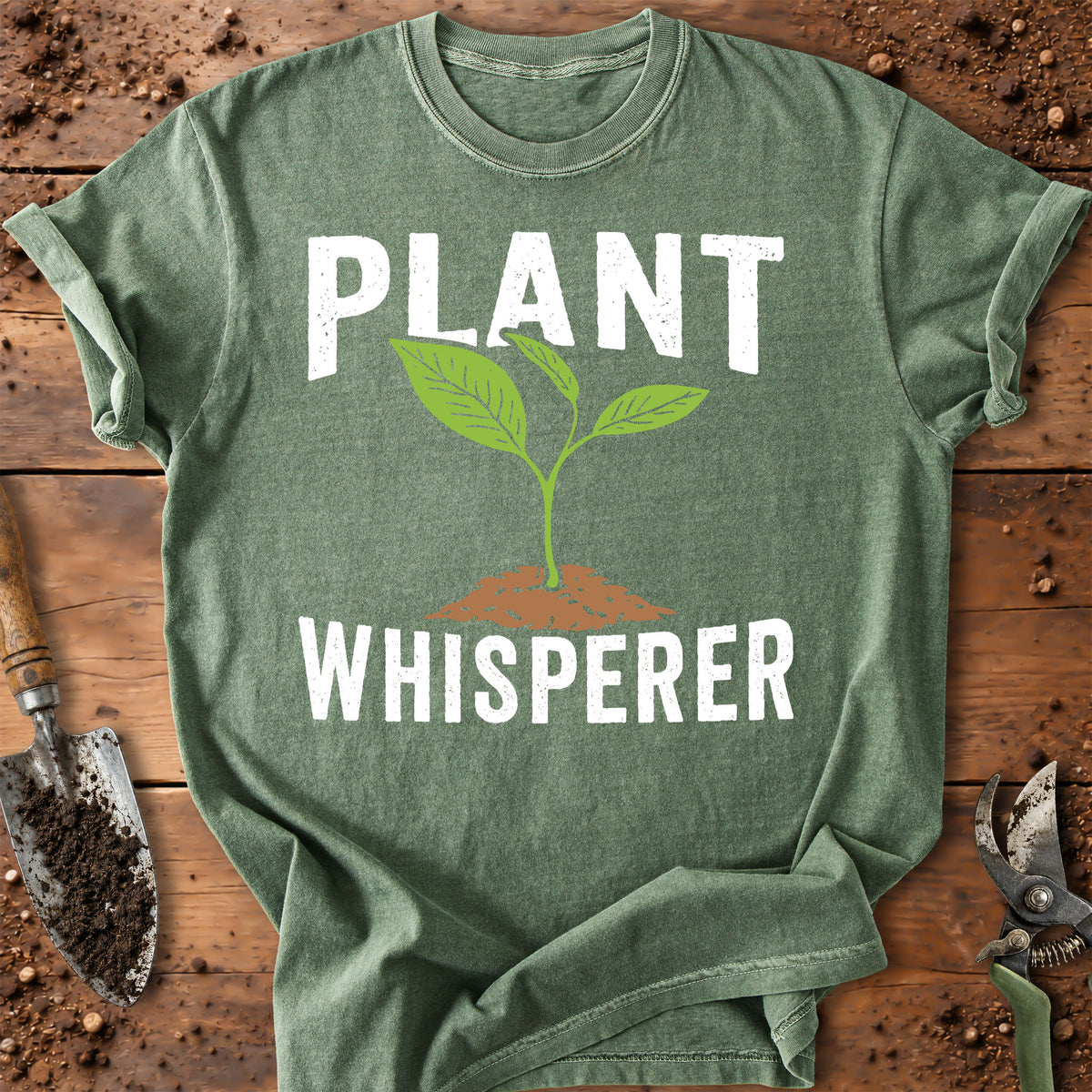 An olive green graphic t-shirt laid flat on a rustic wooden surface with gardening props. The shirt features a white graphic that reads 'PLANT WHISPERER' with a green sprout growing from brown soil in the center.