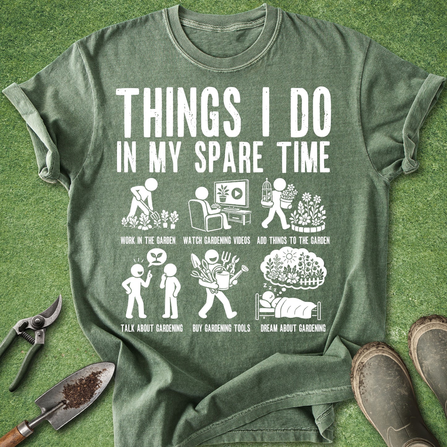A green graphic t-shirt laid flat on a green grass-like surface, surrounded by gardening props including pruning shears, a trowel, and boots. The shirt features a white distressed text and illustration graphic reading 'THINGS I DO IN MY SPARE TIME' with gardening-themed doodles and labels.