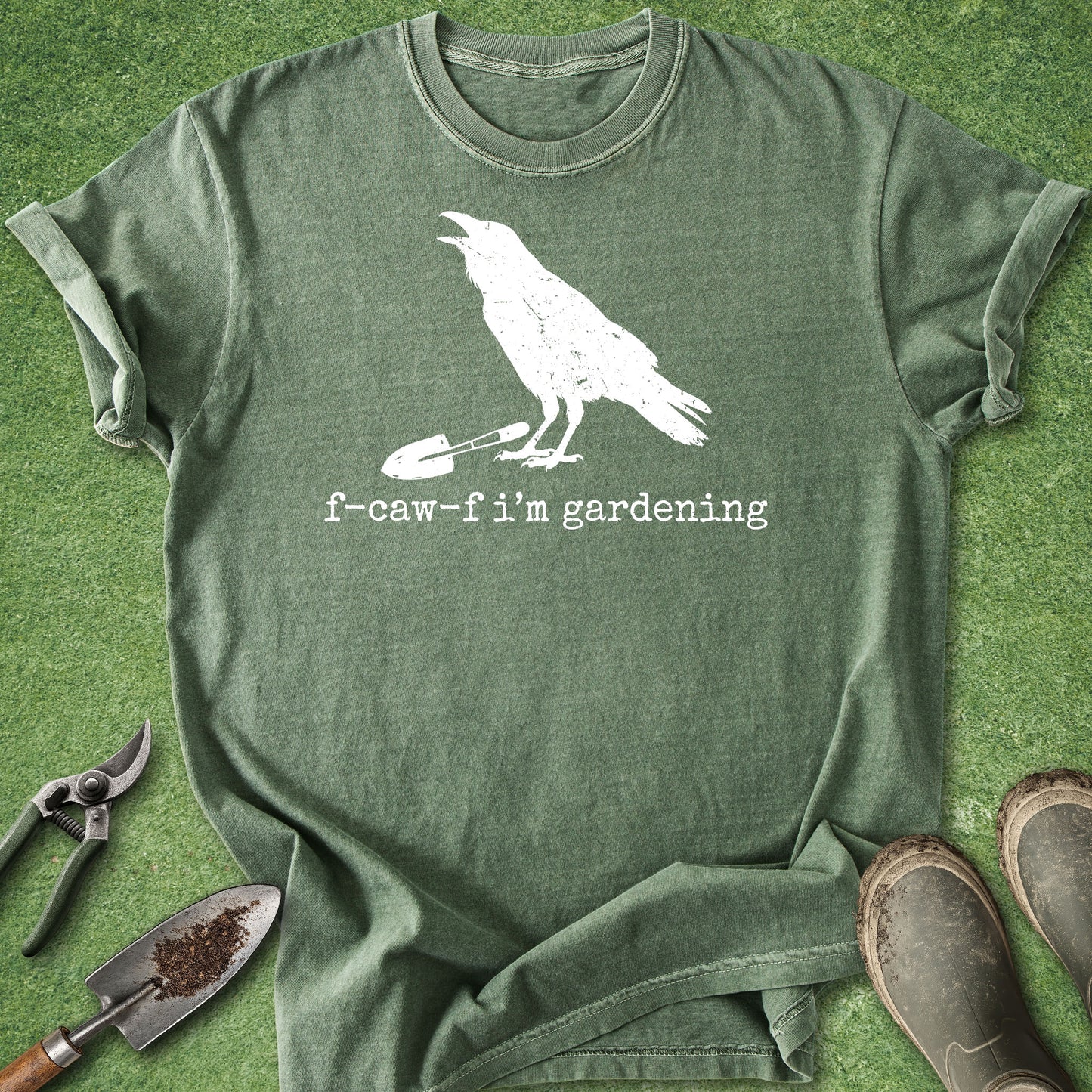 An olive green graphic t-shirt laid flat on a green grass-like surface, featuring a white distressed print of a raven holding a trowel with the text 'f-caw-f i'm gardening'. Gardening props including pruning shears, a trowel with soil, and muddy boots are arranged around the shirt.