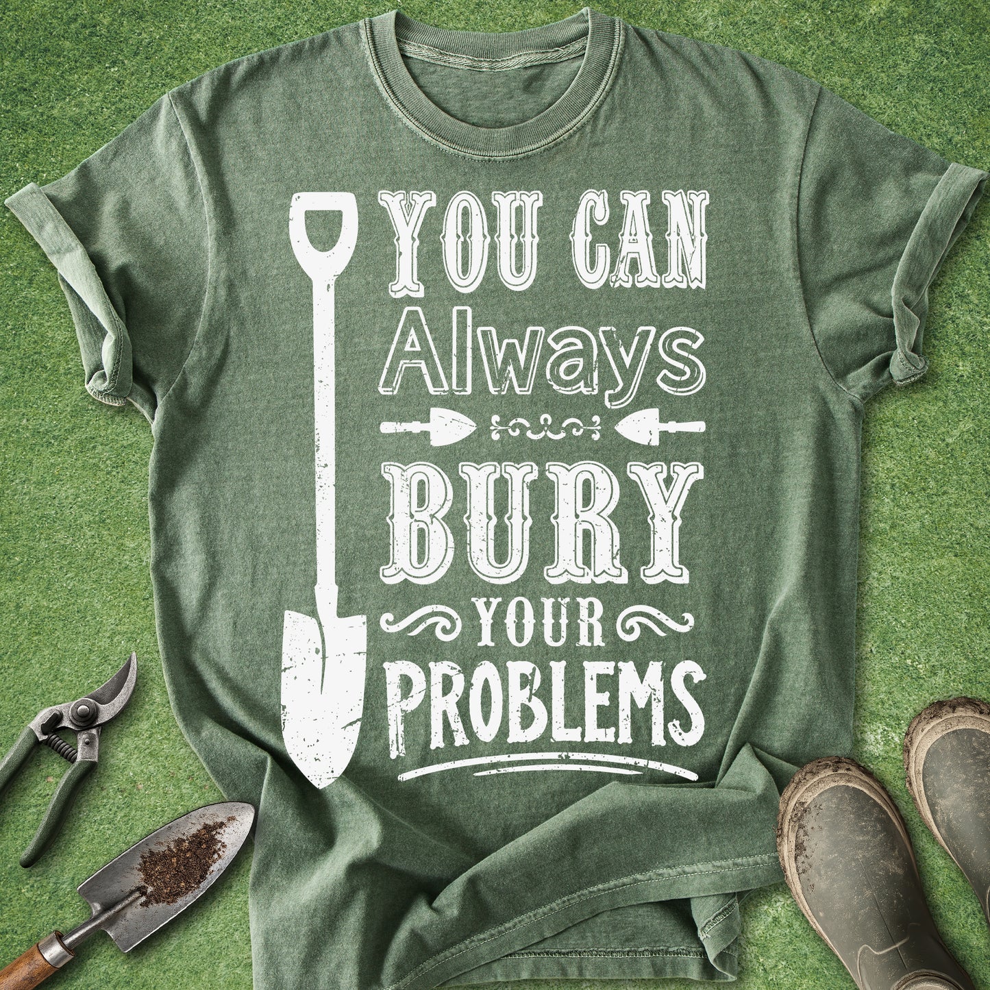 An olive green graphic t-shirt laid flat on a green grass-like surface, featuring a white distressed print that reads 'YOU CAN Always BURY YOUR PROBLEMS' with a shovel illustration. Gardening props including pruning shears, a trowel, and boots are placed around the shirt.