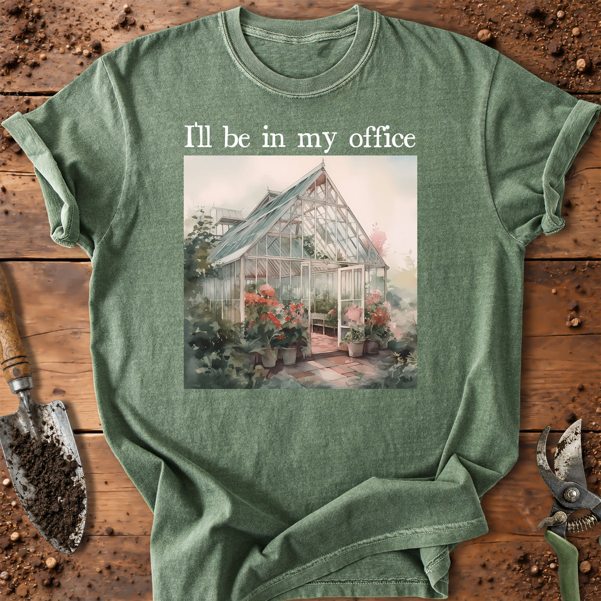 A sage green graphic t-shirt laid flat on a rustic wooden surface with gardening tools. The shirt features a printed illustration of a greenhouse surrounded by flowers and the text 'I'll be in my office' in white above the graphic.
