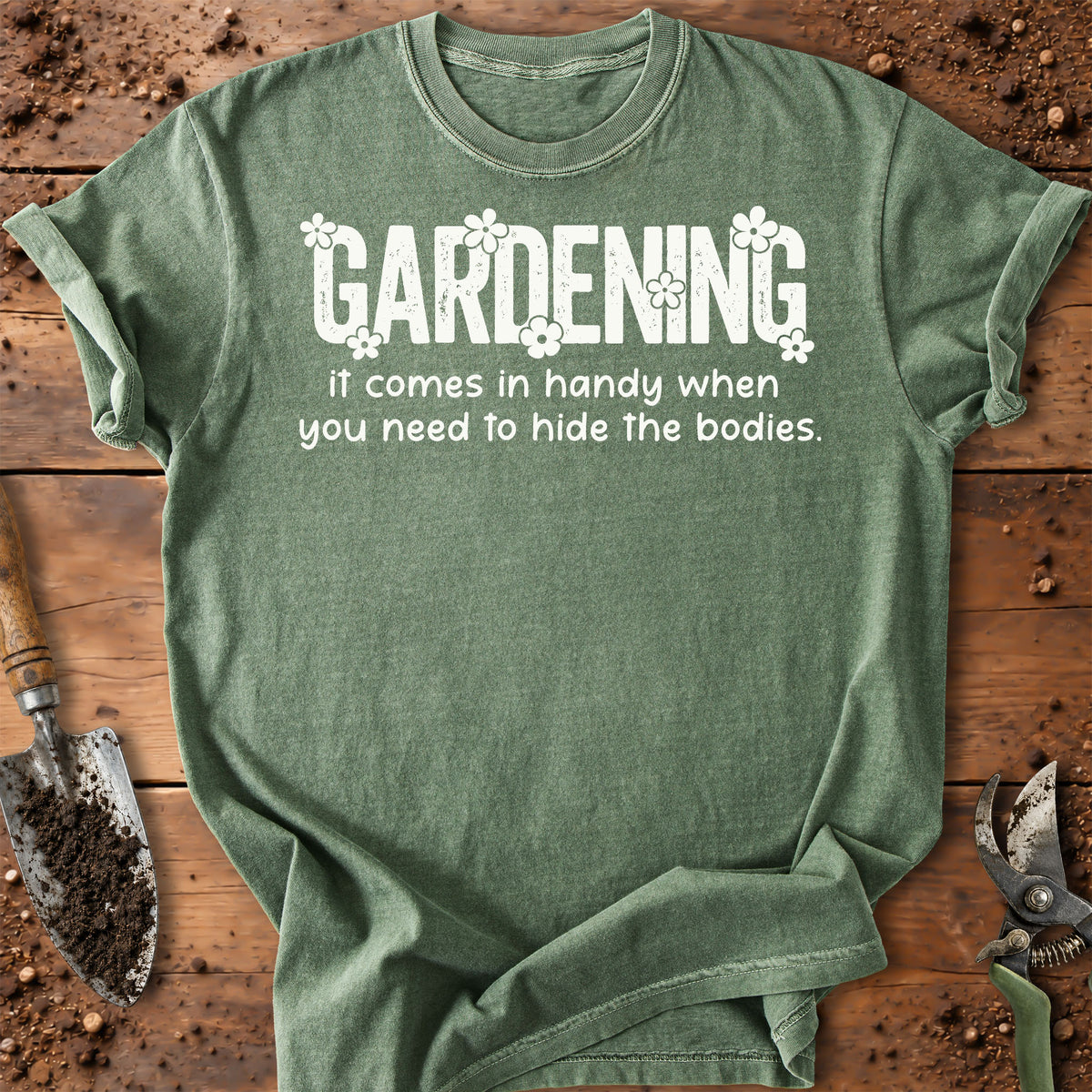 A heathered sage green t-shirt with a white graphic text that reads 'GARDENING it comes in handy when you need to hide the bodies.' surrounded by small white flowers.