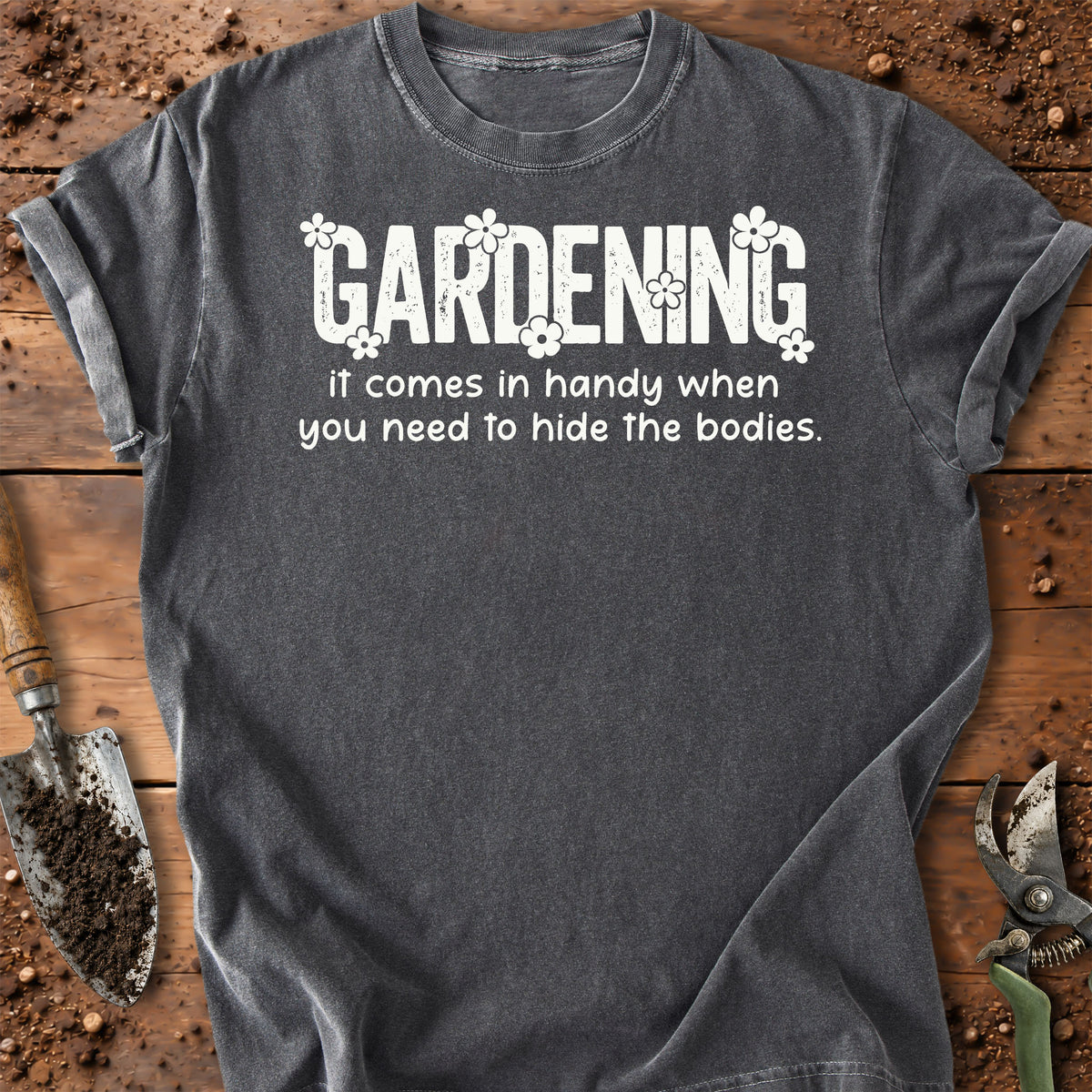 A pepper color short-sleeve t-shirt with a white graphic text that reads 'GARDENING It comes in handy when you need to hide the bodies.' styled in a flat-lay on a rustic wooden background with gardening 