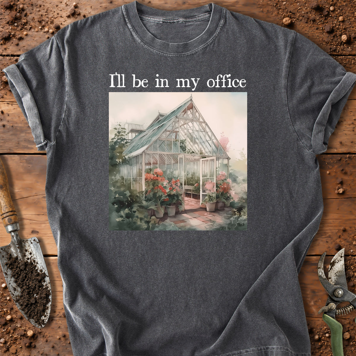 A dark gray graphic t-shirt laid flat on a rustic wooden surface with gardening tools. The shirt features a square graphic of a greenhouse surrounded by flowers, with the text 'I'll be in my office' printed above it.