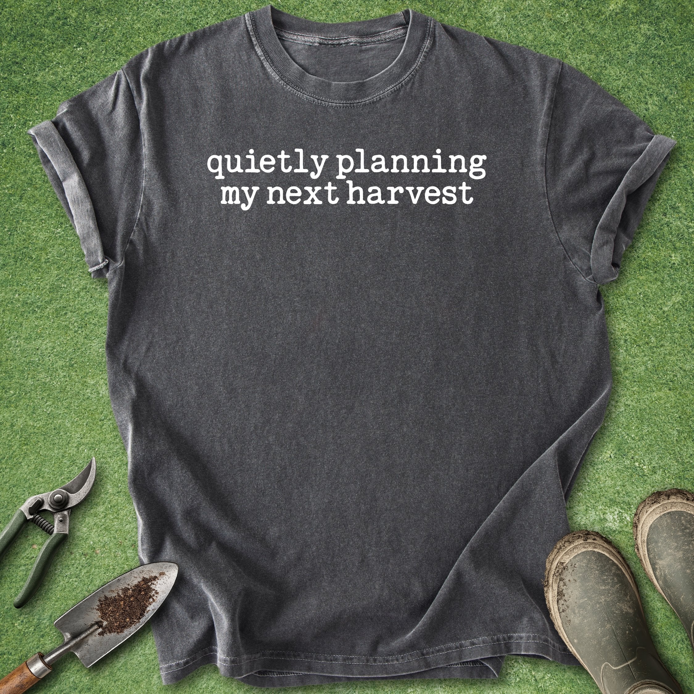 Black t-shirt with 'quietly planning my next harvest' text on a green grass background with gardening tools.