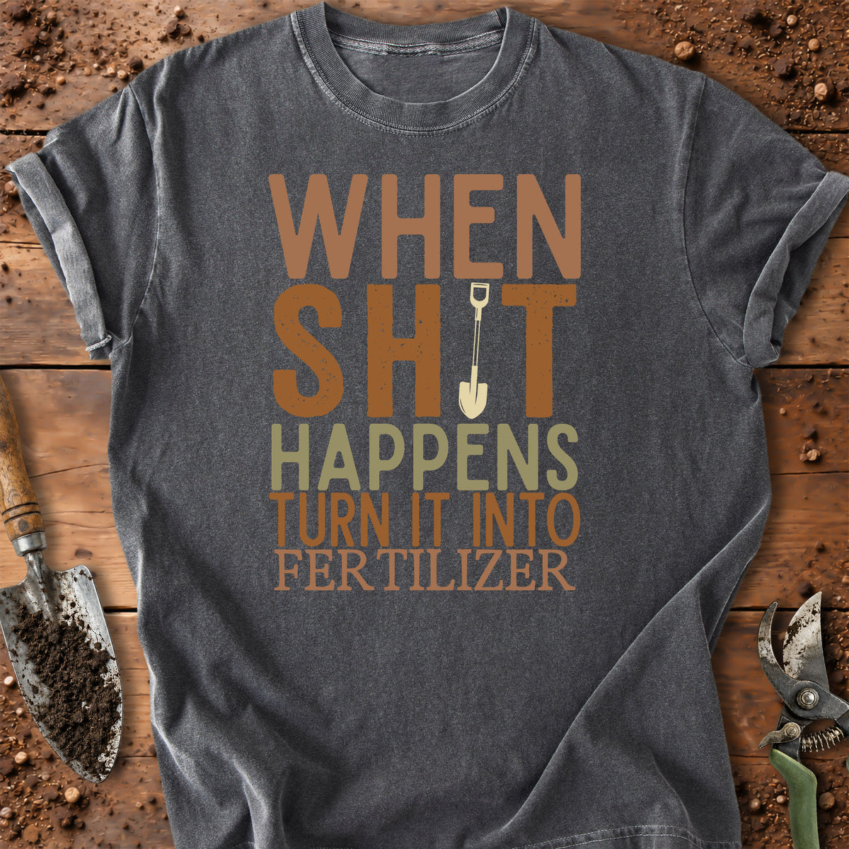 A dark gray graphic t-shirt laid flat on a rustic wooden surface with gardening tools. The shirt features a humorous text design in brown, beige, and light green colors: 'WHEN SHIT HAPPENS TURN IT INTO FER TILIZER' with a small shovel graphic.