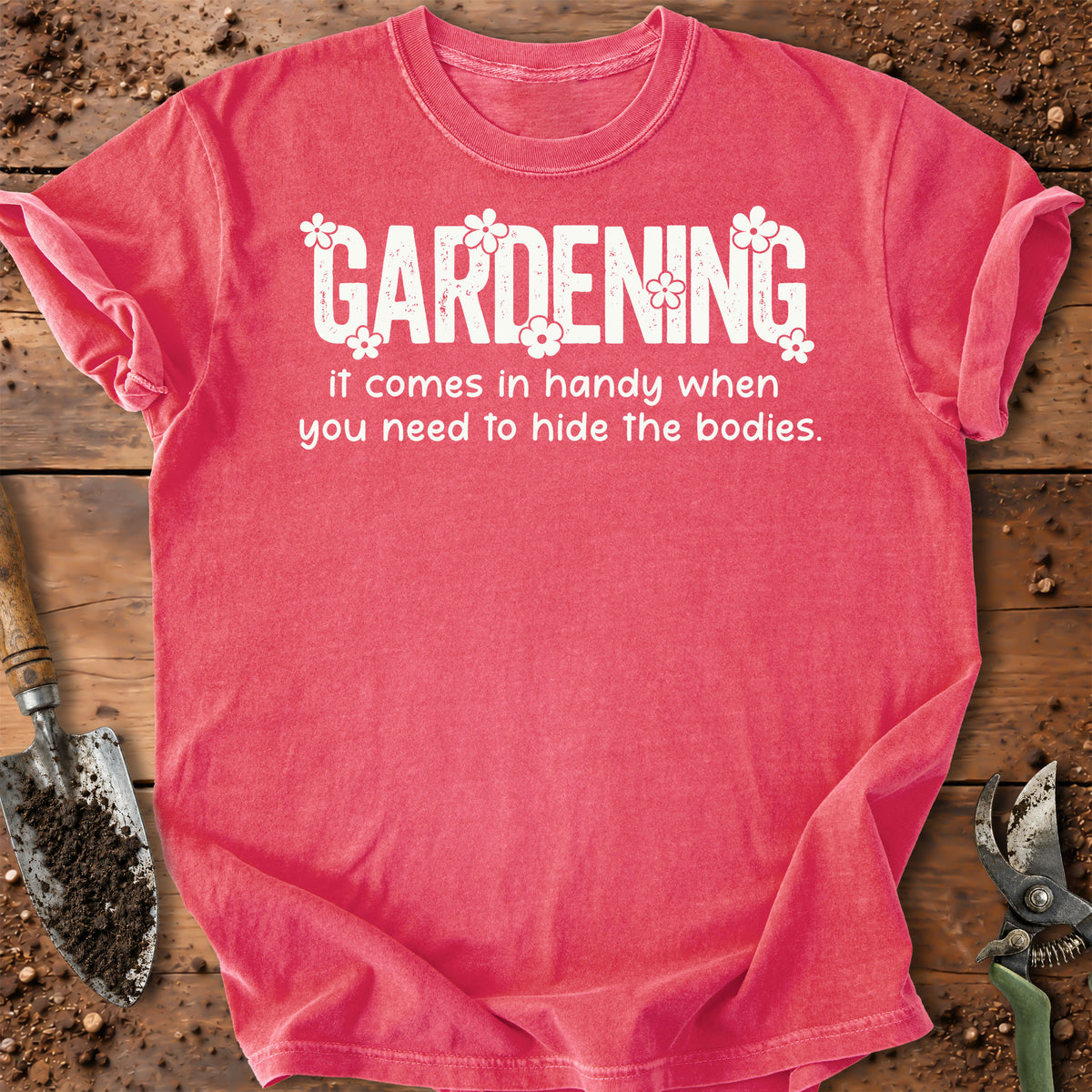A watermelon color short-sleeve t-shirt with a white graphic text that reads 'GARDENING It comes in handy when you need to hide the bodies.' styled in a flat-lay on a rustic wooden background with gardening 