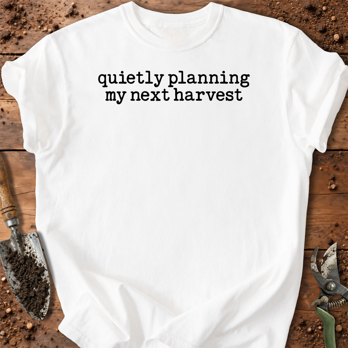 A white short-sleeve crewneck t-shirt with the black printed text 'quietly planning my next harvest' laid flat on a rustic wooden surface with gardening tools like a trowel and pruning shears.