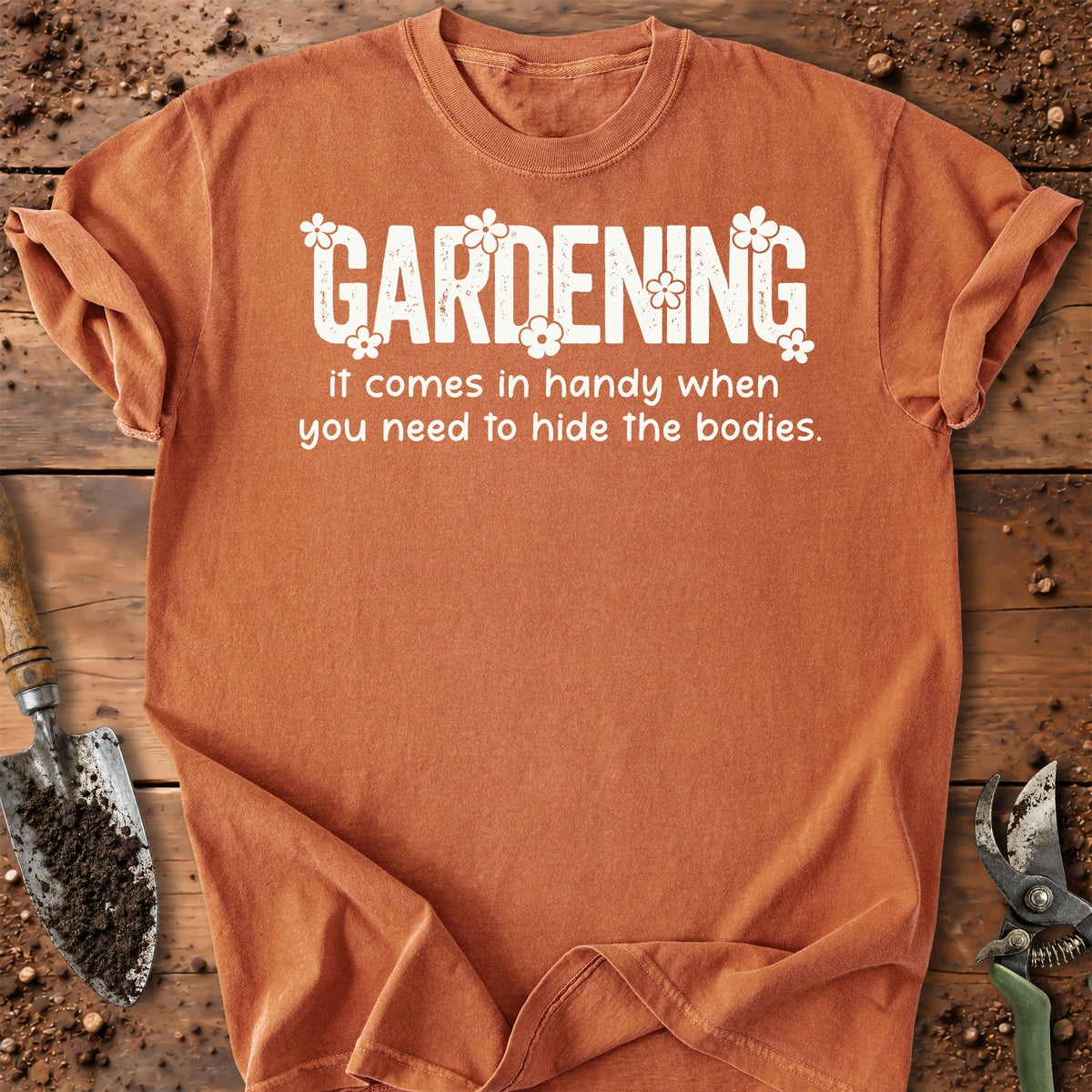 A yam color short-sleeve t-shirt with a white graphic text that reads 'GARDENING It comes in handy when you need to hide the bodies.' styled in a flat-lay on a rustic wooden background with gardening 
