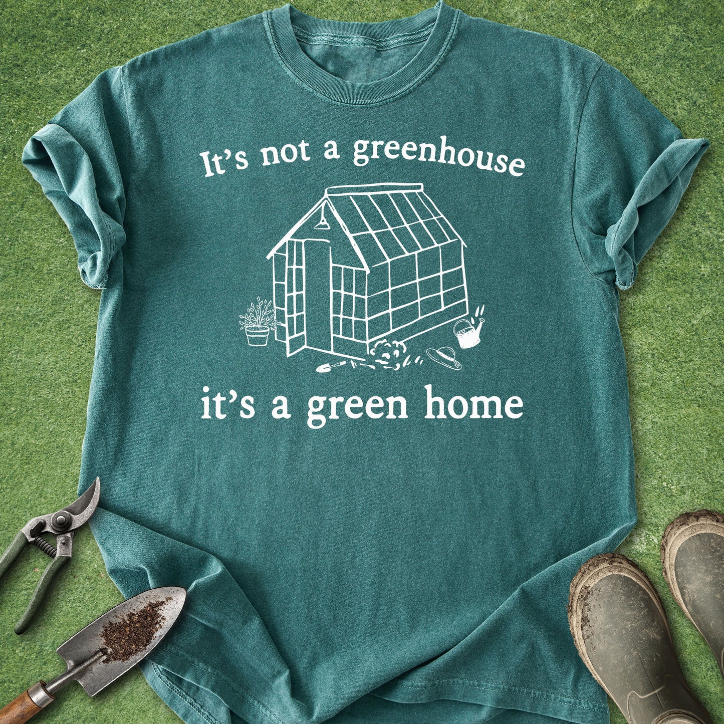 A teal short-sleeve graphic T-shirt laid flat on green grass, with rolled sleeves and a white printed greenhouse illustration and the text 'It's not a greenhouse it's a green home.' Gardening tools and muddy boots are placed around the shirt as styling props.