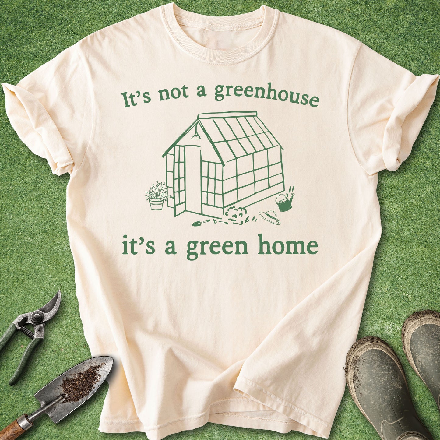 A cream-colored short-sleeve t-shirt with a green graphic of a greenhouse and the text 'It's not a greenhouse it's a green home', laid flat on a green grass-like surface with gardening tools and shoes around it.