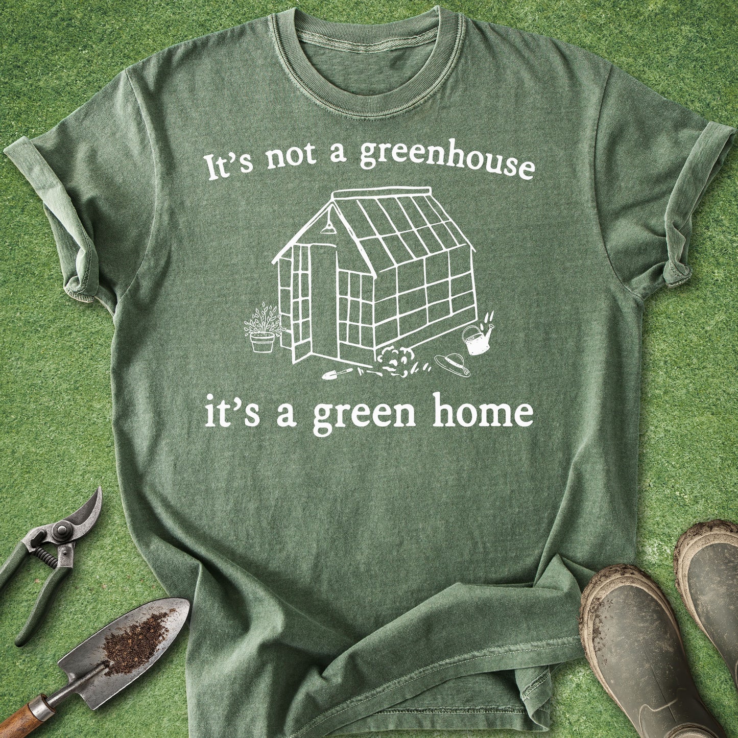 An olive green graphic t-shirt laid flat on a green grass-like surface, surrounded by gardening props including pruning shears, a trowel, and boots. The shirt features a white line drawing of a greenhouse with the text 'It's not a greenhouse it's a green home'.