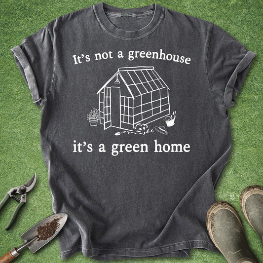 A dark charcoal gray graphic t-shirt laid flat on green grass, featuring a white line art drawing of a greenhouse and the text 'It's not a greenhouse it's a green home'. Gardening props including pruning shears, a trowel, and boots are visible around the shirt.