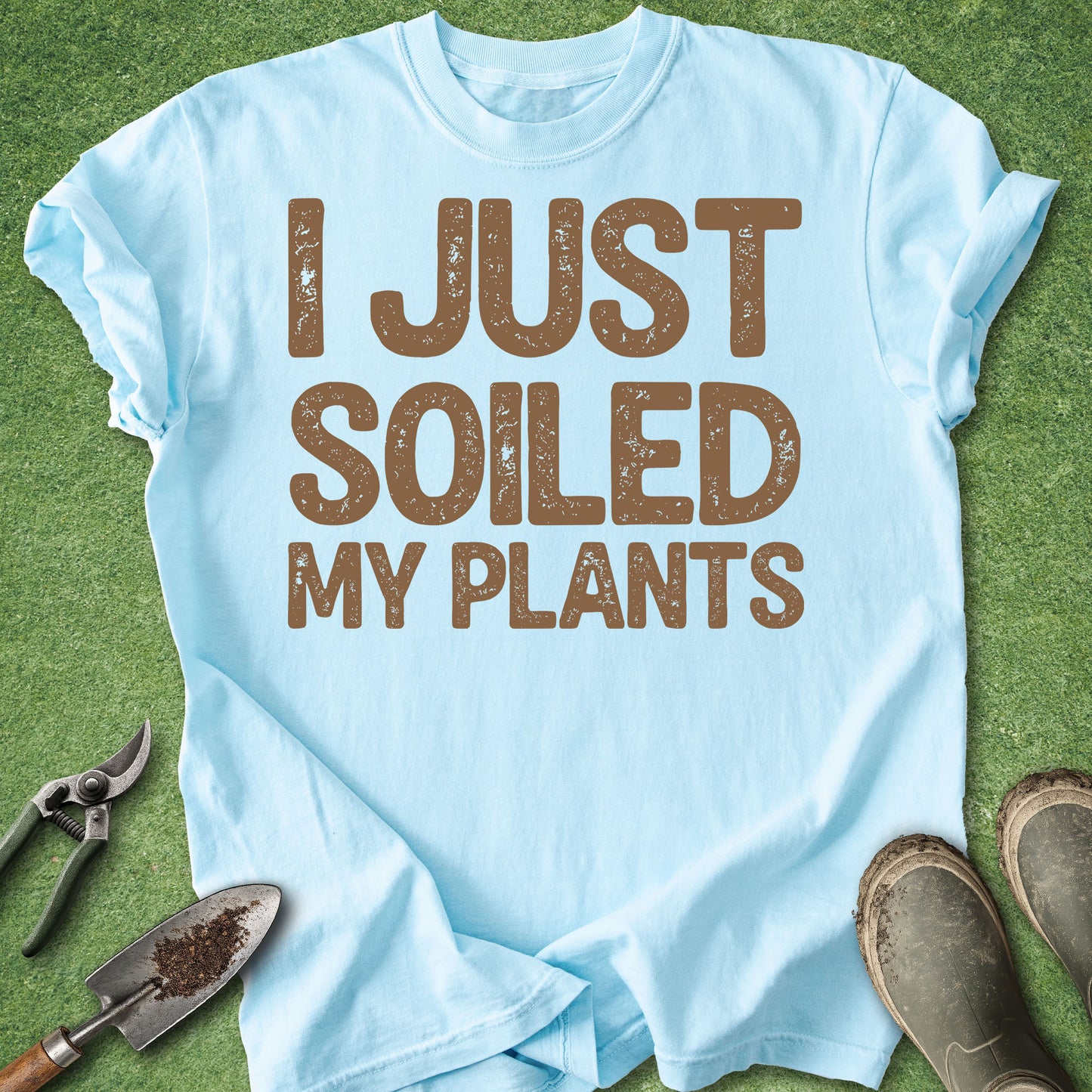 Light blue t-shirt with 'I JUST SOILED MY PLANTS' text on a grass background with gardening tools.