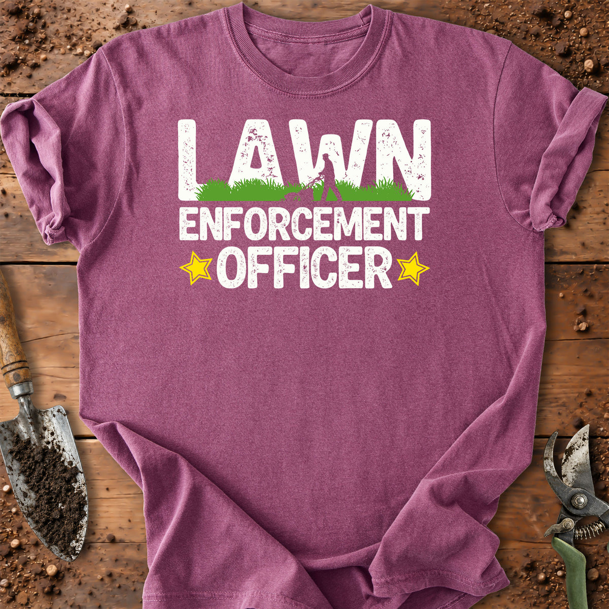 A heathered purple short-sleeve t-shirt laid flat on a rustic wooden surface with gardening props like a trowel and pruning shears. The shirt features a graphic print that reads 'LAWN ENFORCEMENT OFFICER' in white distressed letters, with green grass under the word 'LAWN' and yellow star accents.