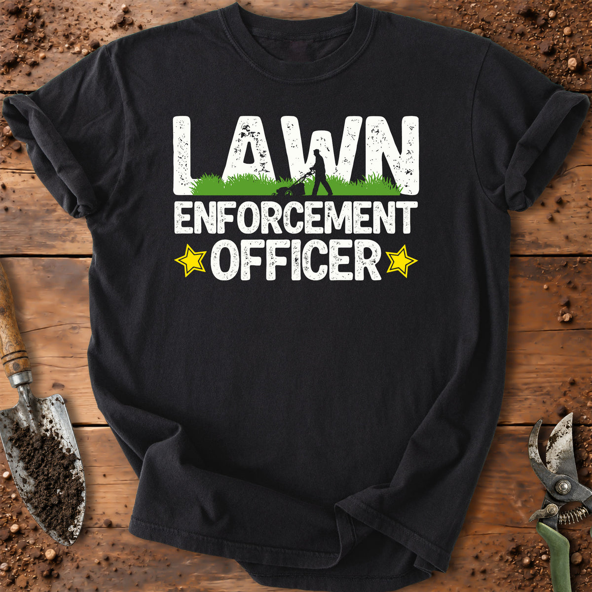 A black short-sleeve t-shirt laid flat on a rustic wooden surface with soil and gardening tools around it. The shirt has a large front graphic reading 'LAWN ENFORCEMENT OFFICER' in white distressed letters, with a small green grass area, a silhouette of a person, and two yellow stars.