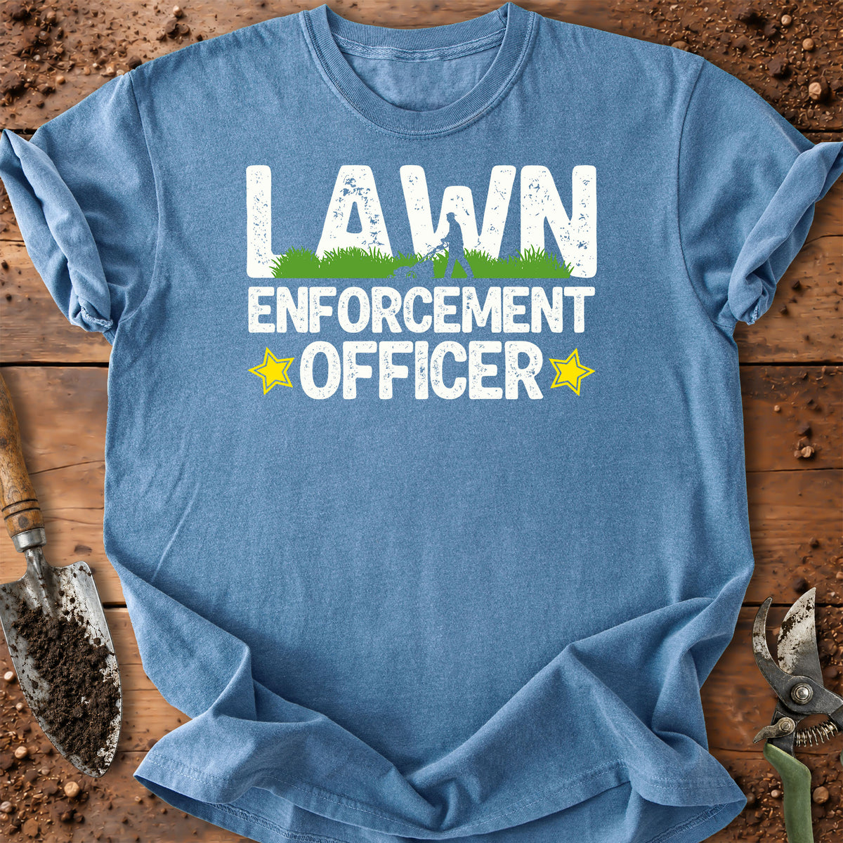 A light blue crewneck t-shirt laid flat on a rustic wooden surface with gardening tools. The shirt features a distressed white graphic that reads 'LAWN ENFORCEMENT OFFICER' with a small green grass graphic and yellow stars.