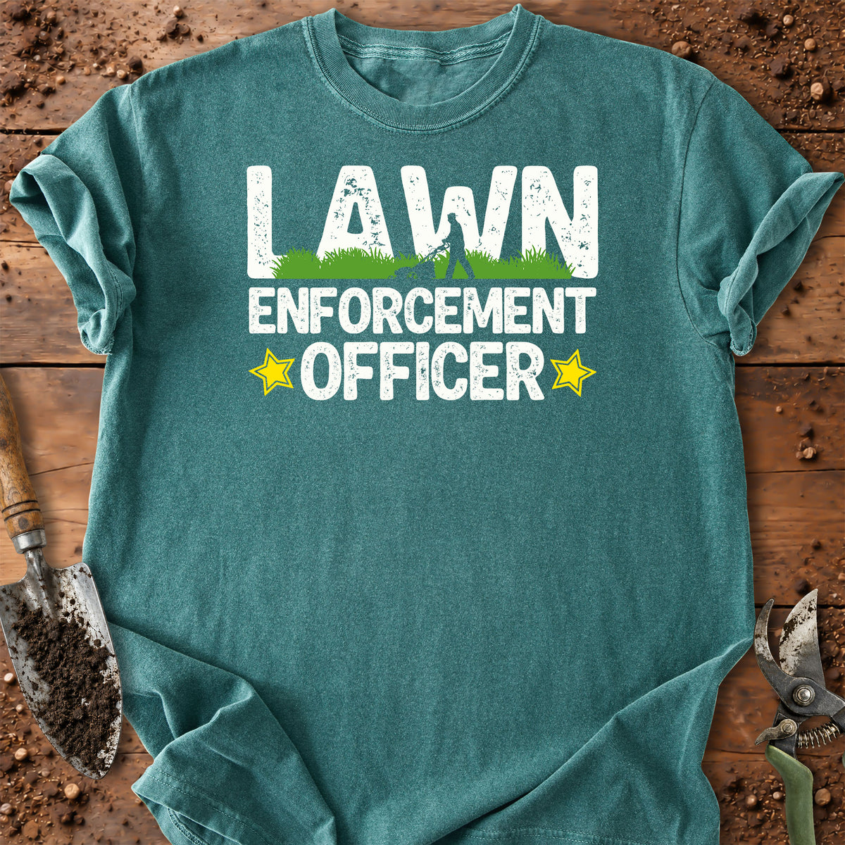 A teal short-sleeve crewneck t-shirt laid flat on a rustic wooden surface with gardening props like a trowel and pruning shears. The shirt features a graphic print that reads 'LAWN ENFORCEMENT OFFICER' in distressed white letters, with a small green grass graphic and yellow stars.