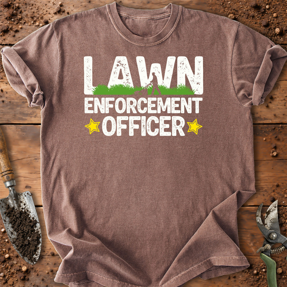A brown short-sleeve crewneck t-shirt laid flat on a rustic wooden surface with gardening props. The shirt features a graphic print that reads 'LAWN ENFORCEMENT OFFICER' in white distressed letters, with a small green grass graphic inside the letter 'A' and two yellow stars on either side.