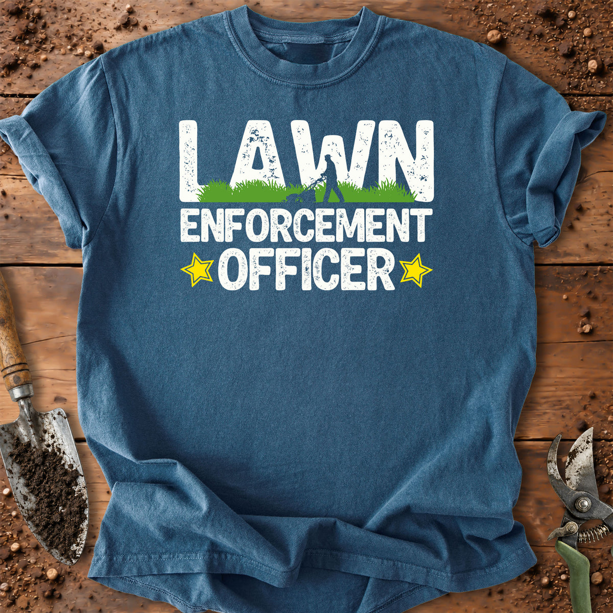 A blue crewneck t-shirt laid flat on a rustic wooden surface with gardening tools around it. The shirt features a graphic print that reads 'LAWN ENFORCEMENT OFFICER' in white, with a small green grass graphic inside the 'A' in 'LAWN' and two yellow stars on either side of 'OFFICER'.