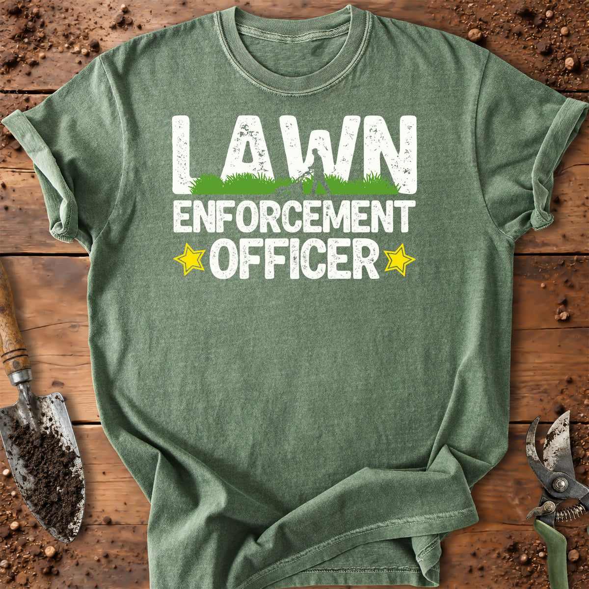 A sage green graphic t-shirt laid flat on a rustic wooden background with gardening props like a trowel and pruning shears. The shirt features a distressed white graphic that reads 'LAWN ENFORCEMENT OFFICER' with a small green grass graphic and yellow stars.