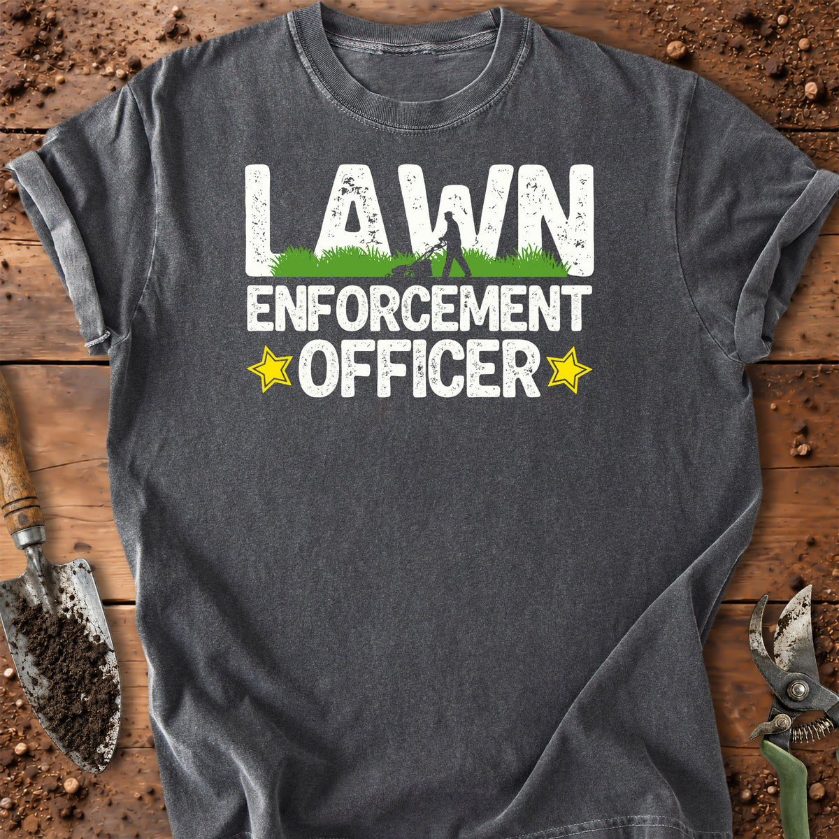 A dark gray graphic t-shirt laid flat on a rustic wooden surface with gardening props. The shirt features a distressed white graphic that reads 'LAWN ENFORCEMENT OFFICER' with a small green grass graphic and yellow stars.
