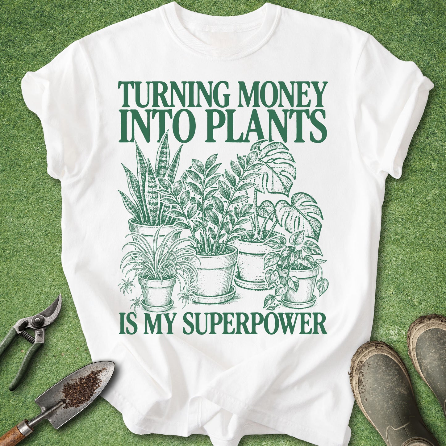 A white short-sleeve graphic T-shirt laid flat on green grass, with dark green text reading 'TURNING MONEY INTO PLANTS IS MY SUPERPOWER' and an illustration of potted plants. Gardening tools and boots are placed around the shirt as styled props.