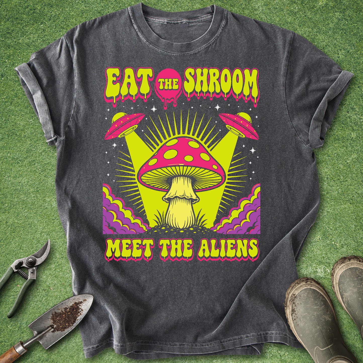A dark gray graphic t-shirt laid flat on green grass, featuring a colorful psychedelic design with a large pink and yellow mushroom, two flying saucers, and the text 'EAT THE SHROOM MEET THE ALIENS'. A trowel, pruning shears, and boots are placed around the shirt as props.