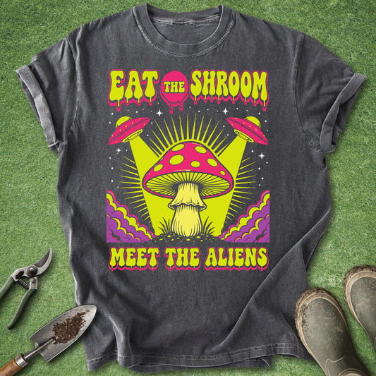 A dark gray graphic t-shirt laid flat on green grass, featuring a colorful psychedelic design with a large pink and yellow mushroom, two flying saucers, and the text 'EAT THE SHROOM MEET THE ALIENS'. A trowel, pruning shears, and boots are placed around the shirt as props.