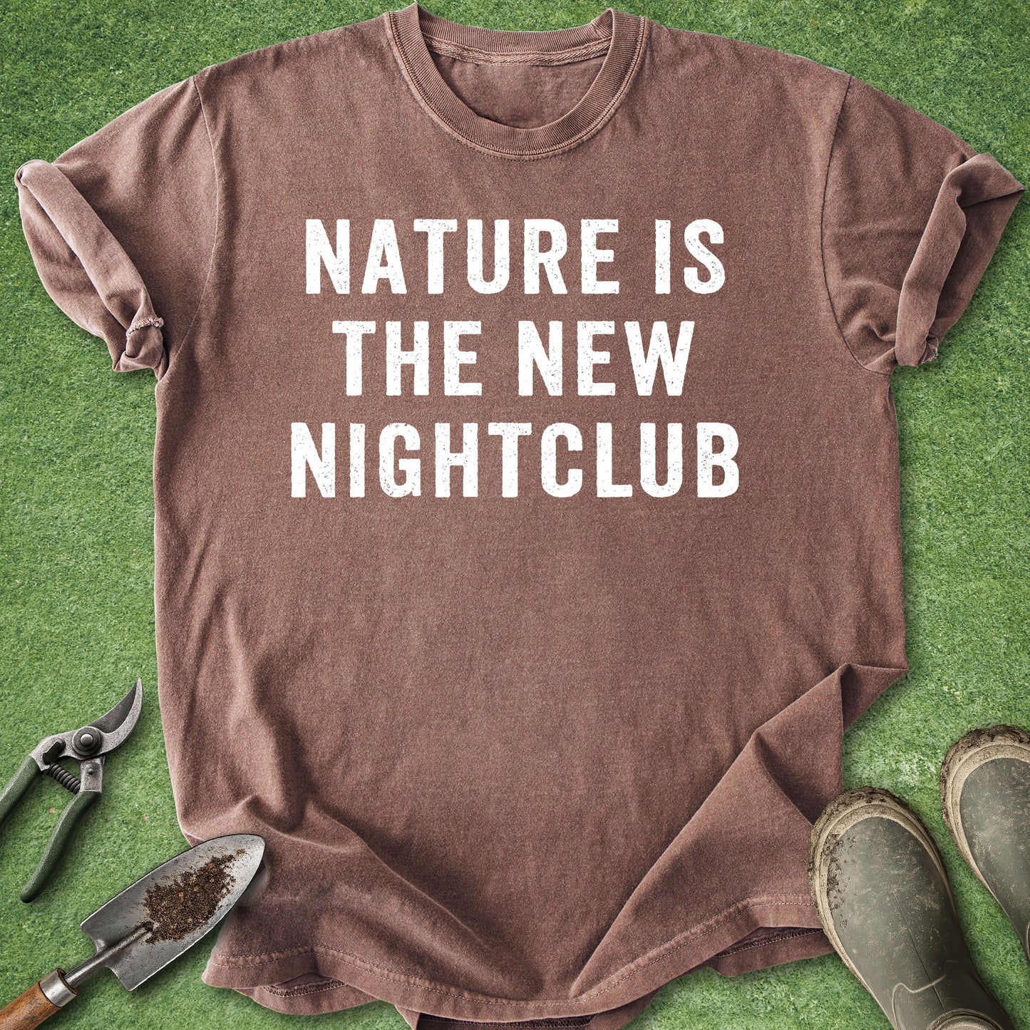 A brown short-sleeve t-shirt with the text 'NATURE IS THE NEW NIGHTCLUB' printed in white capital letters, laid flat on green grass with gardening tools and boots around it.