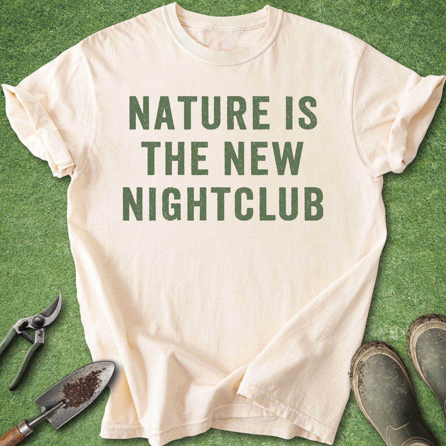 A cream-colored short-sleeve t-shirt with the text 'NATURE IS THE NEW NIGHTCLUB' printed in dark green block letters, laid flat on a green grass background with gardening tools and muddy boots nearby.