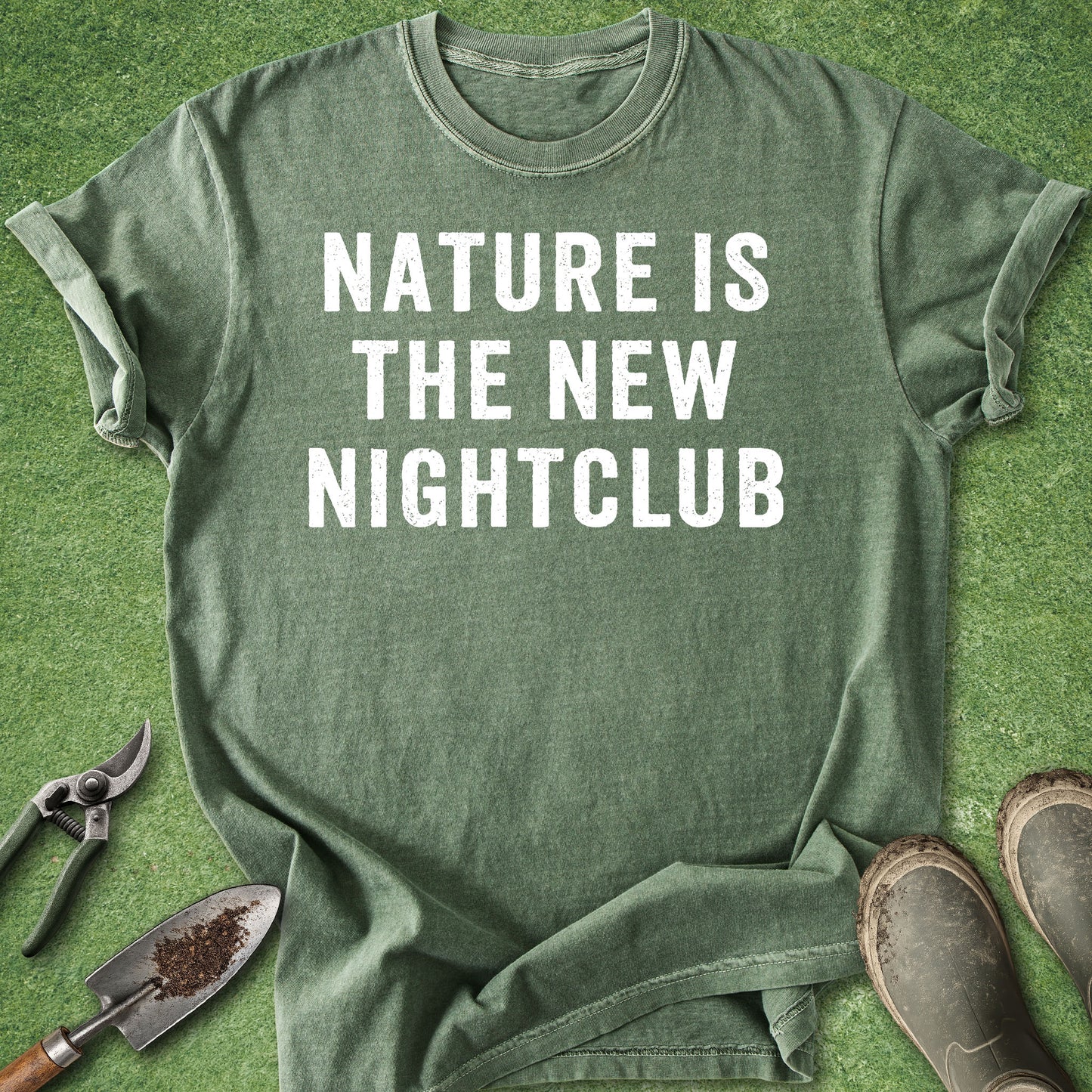 An olive green graphic t-shirt with the text 'NATURE IS THE NEW NIGHTCLUB' in white capital letters, laid flat on green grass with gardening tools (trowel, pruning shears) and work boots as props.